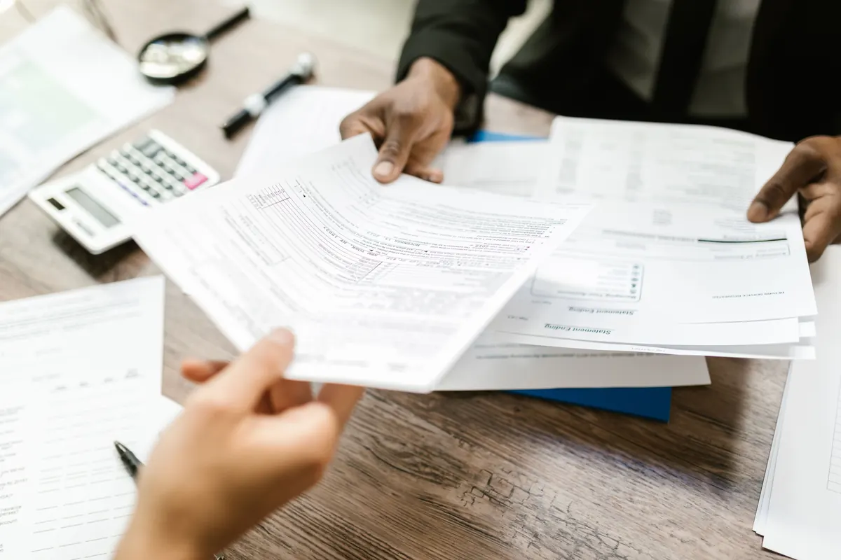 Two people passing printed statements across a wooden desk with a calculator and a magnifying glass