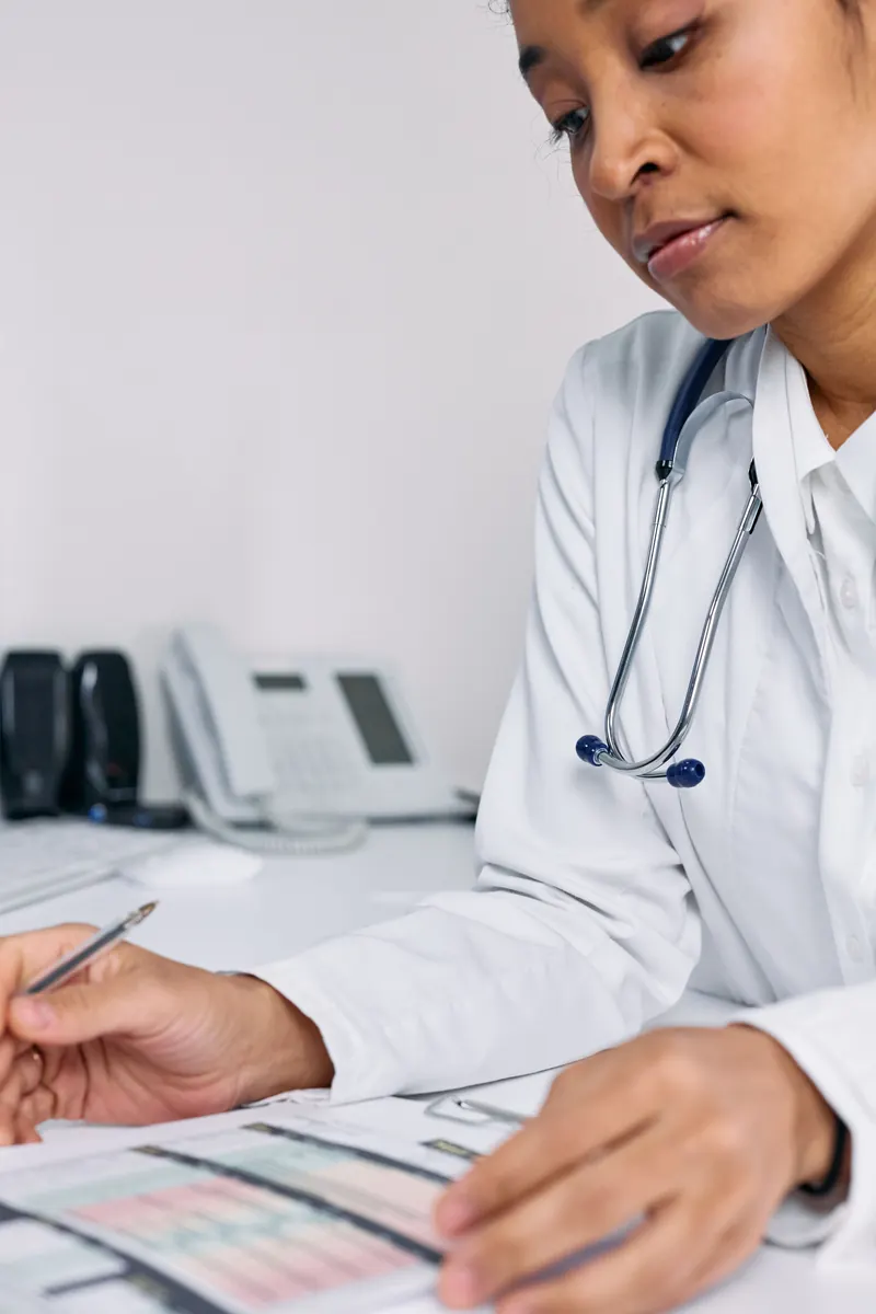 A doctor in a white coat with a stethoscope reading a printed chart, pen in hand
