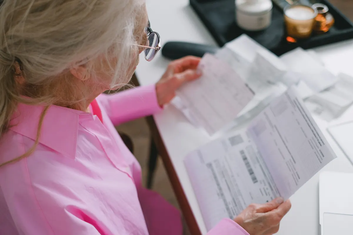 An older woman with long gray hair and glasses in a pink shirt reading printed bills at a table