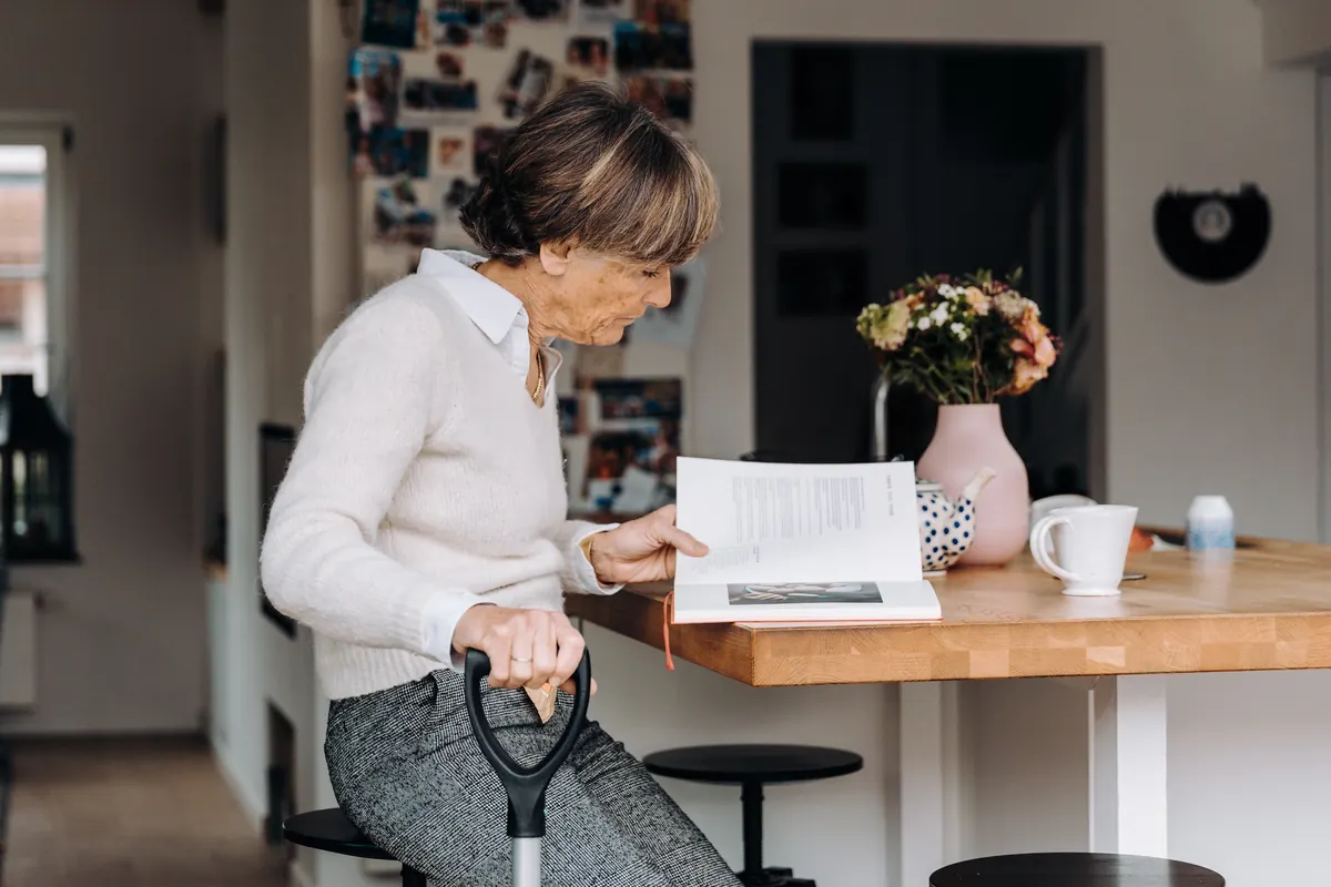 An older woman holding a cane, sitting at a kitchen counter and reading a book beside flowers and a mug