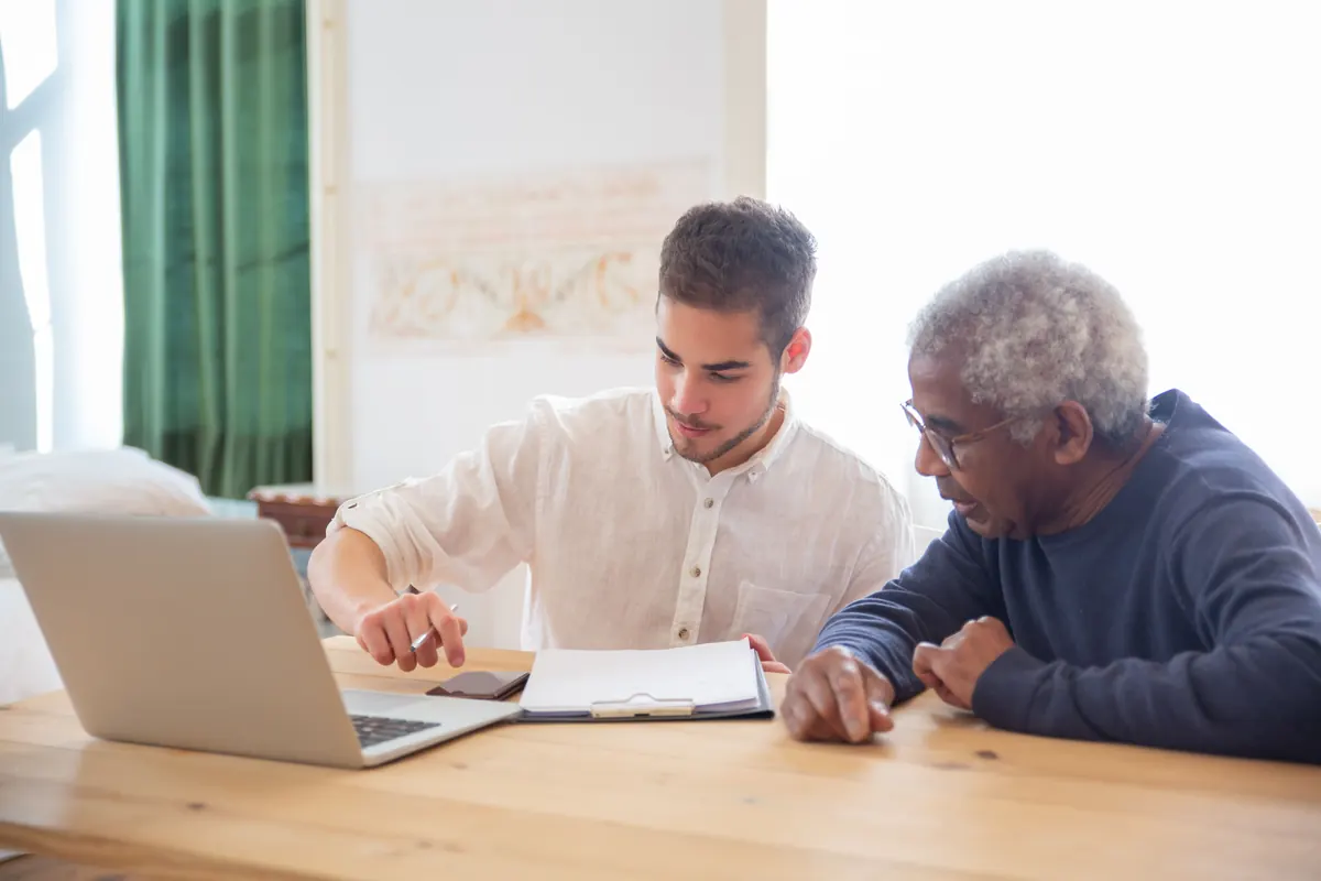 A young man with a pen and an older man with glasses going over papers at a table with a laptop