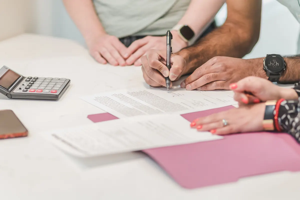 Three people's hands at a white table, one signing papers on a pink folder beside a calculator and phone
