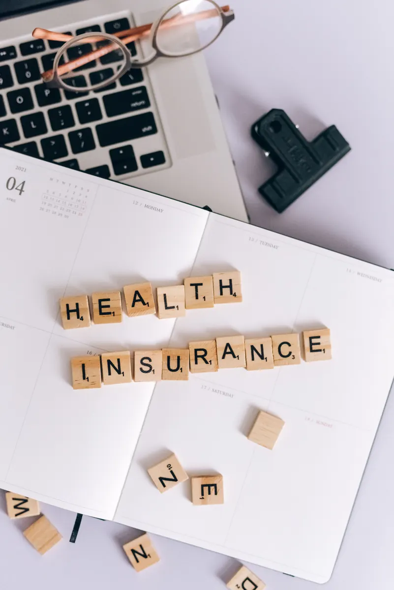 Wooden letter tiles spelling HEALTH INSURANCE on an open planner, with a laptop and glasses nearby
