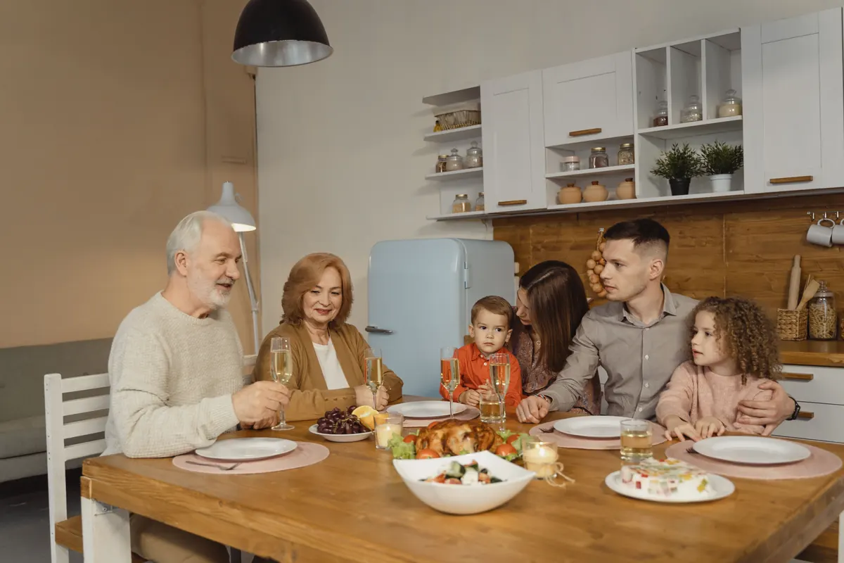 Three generations of a family seated around a kitchen table set for a holiday meal with a roast