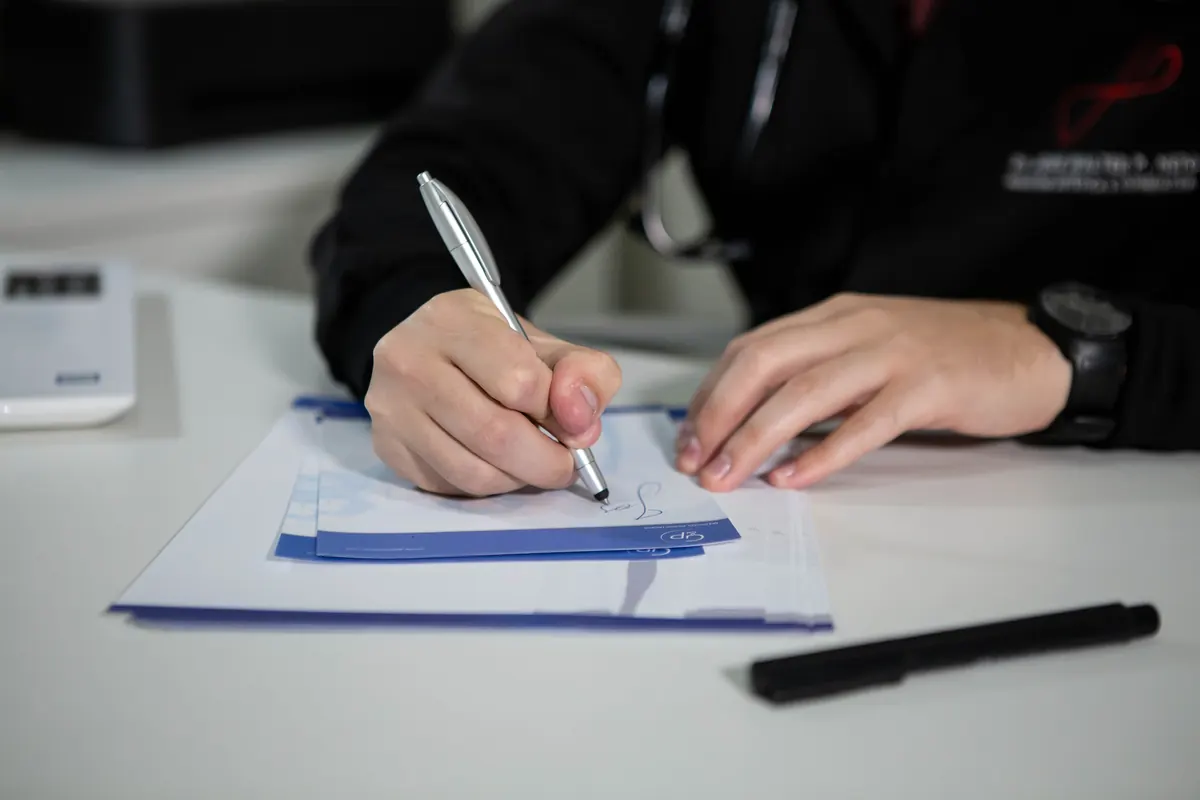 Close-up of a person in a dark jacket writing on a blue-edged notepad on a white desk