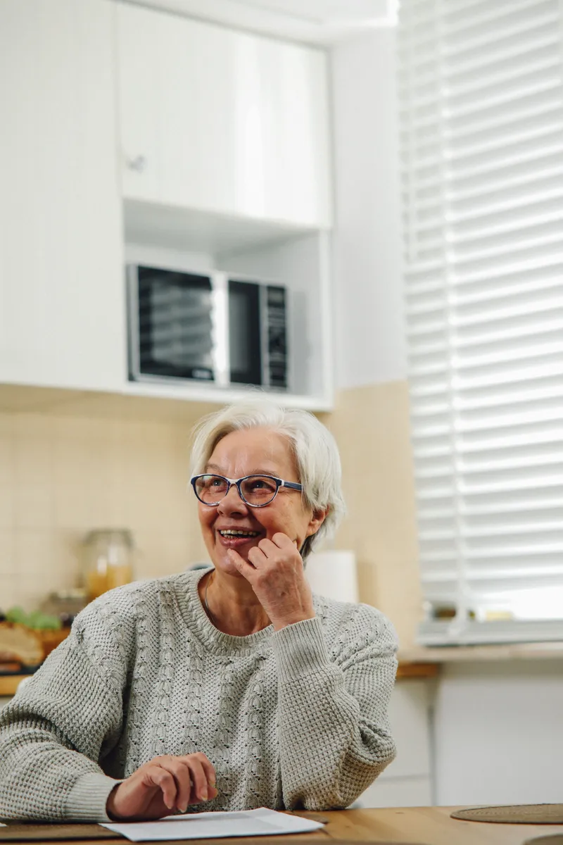 A smiling older woman with white hair and glasses sitting at her kitchen table with a sheet of paper