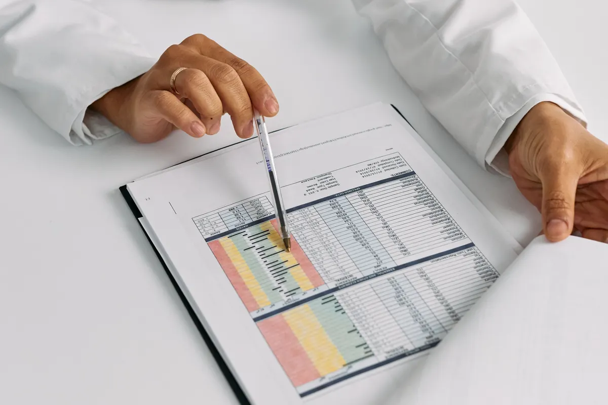 A hand in a white sleeve pointing a pen at a color-coded printed report on a white table