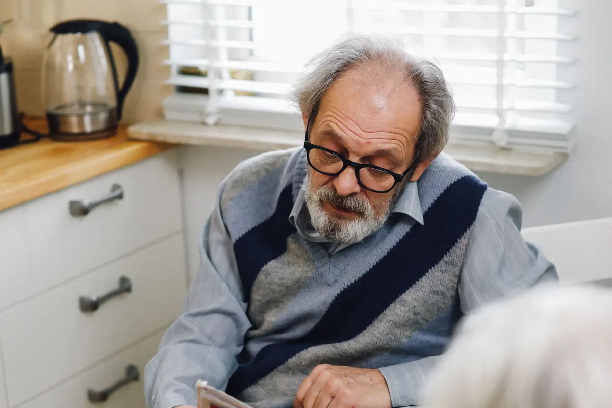 An older man with a gray beard and glasses reading at a kitchen table by a window with blinds