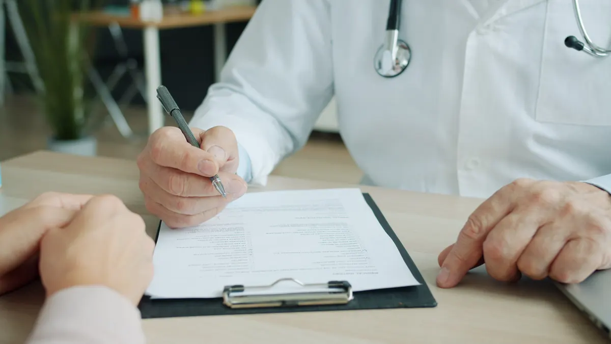 A doctor in a white coat with a stethoscope writing on a clipboard form across a desk from a patient