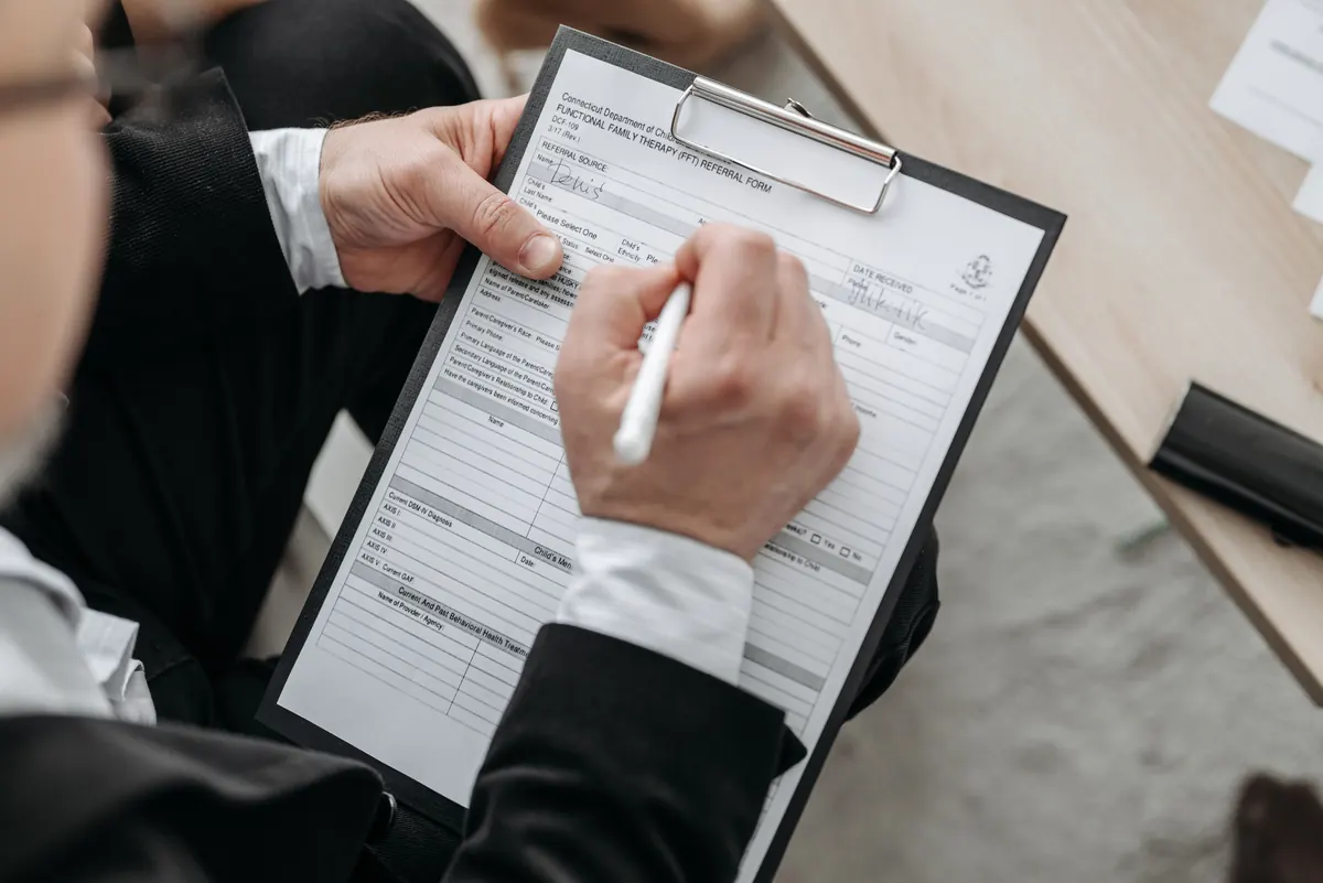 A man in a dark suit filling out a printed form on a clipboard with a white pen