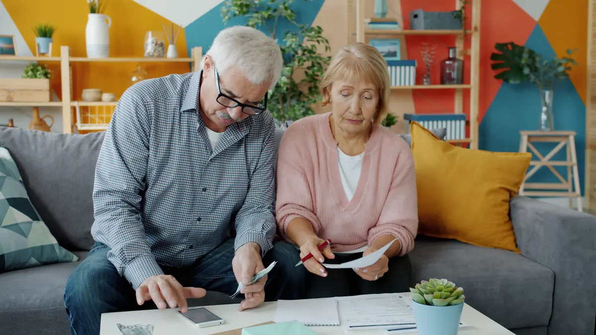 An older man with glasses counting cash beside an older woman holding a letter and a pen on a sofa