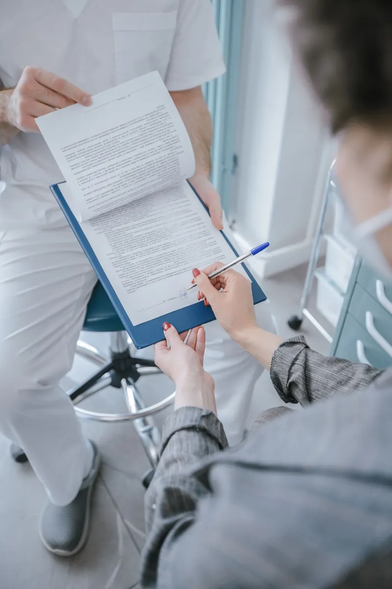 A patient signing a printed form on a blue clipboard held by a health worker dressed in white