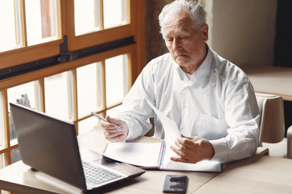 An older man with white hair in a white shirt reading notes at a table by a window, a laptop open