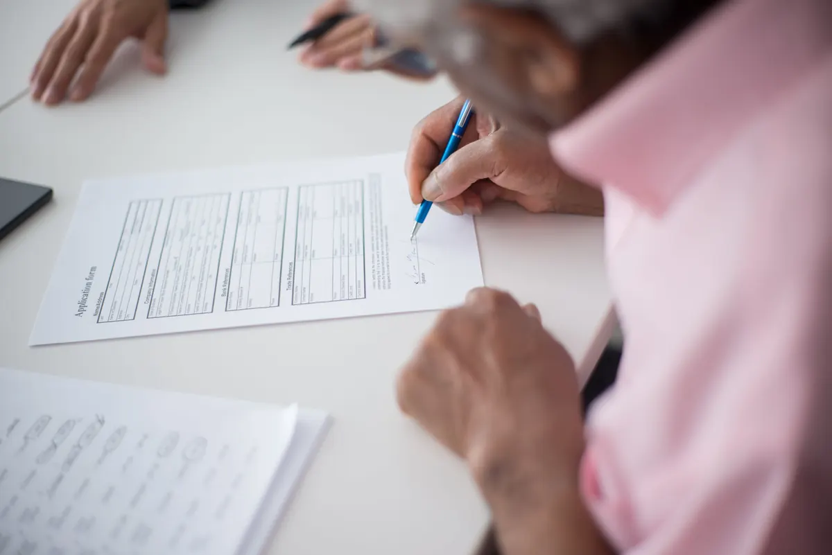 An older man in a pink shirt signing a printed application form at a white table