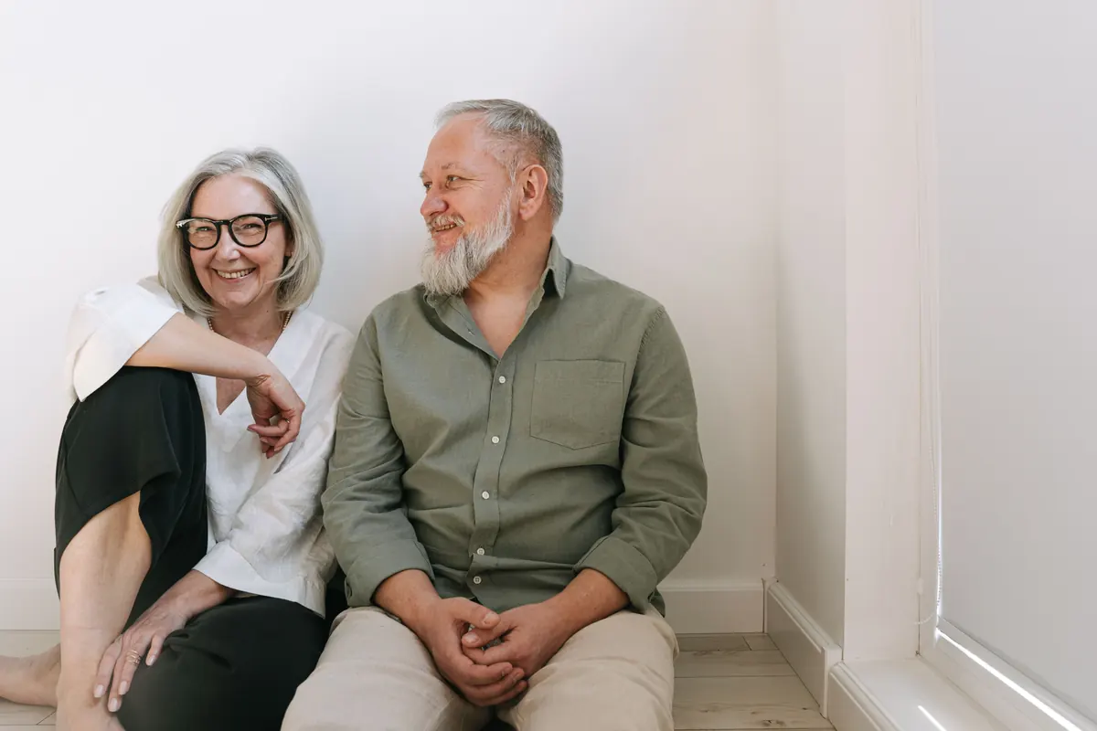 A smiling older couple sitting side by side on the floor against a white wall