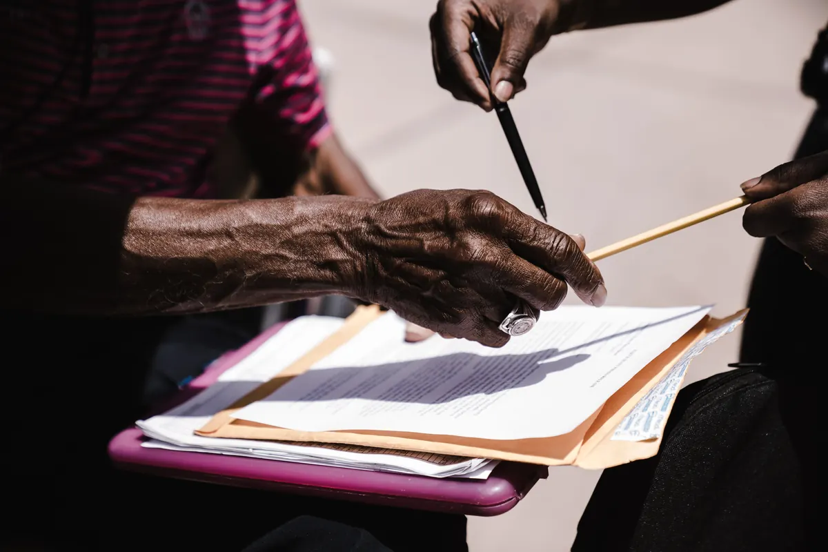 An older person's hand resting on papers in a folder while another person points to them with a pen