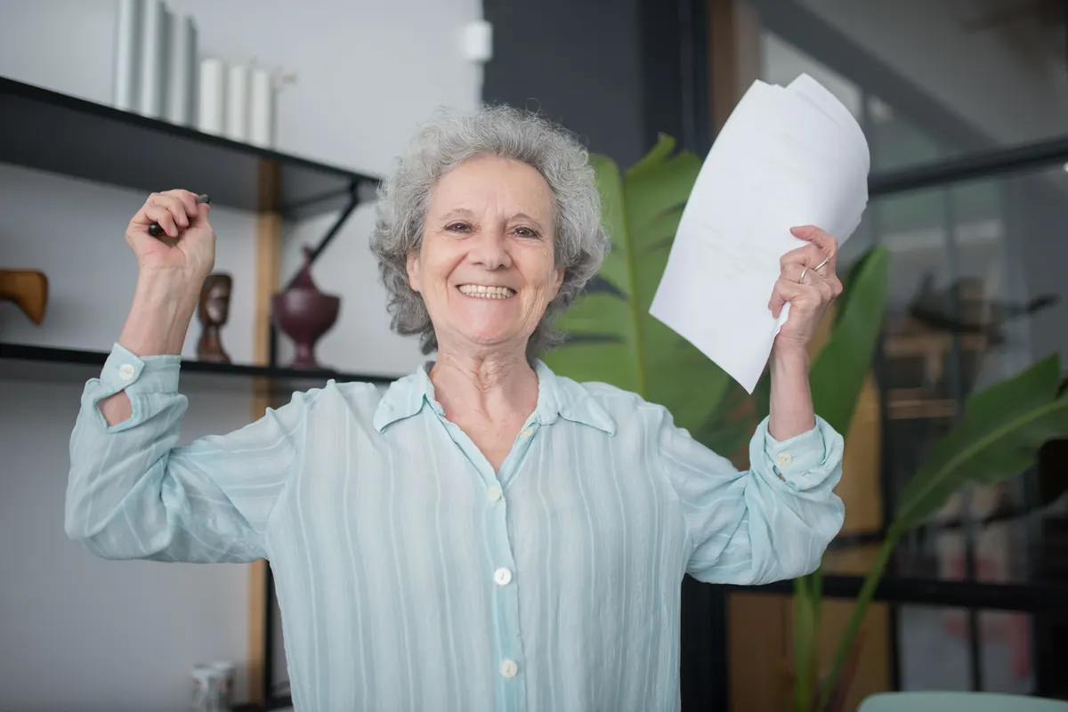 A smiling older woman with gray curly hair raising both arms, holding papers in one hand
