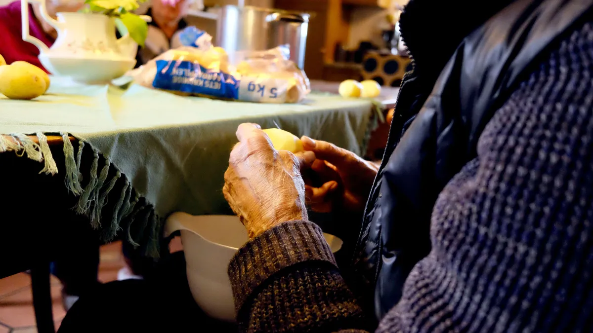 Close-up of an older person's hands peeling a potato over a bowl beside a kitchen table
