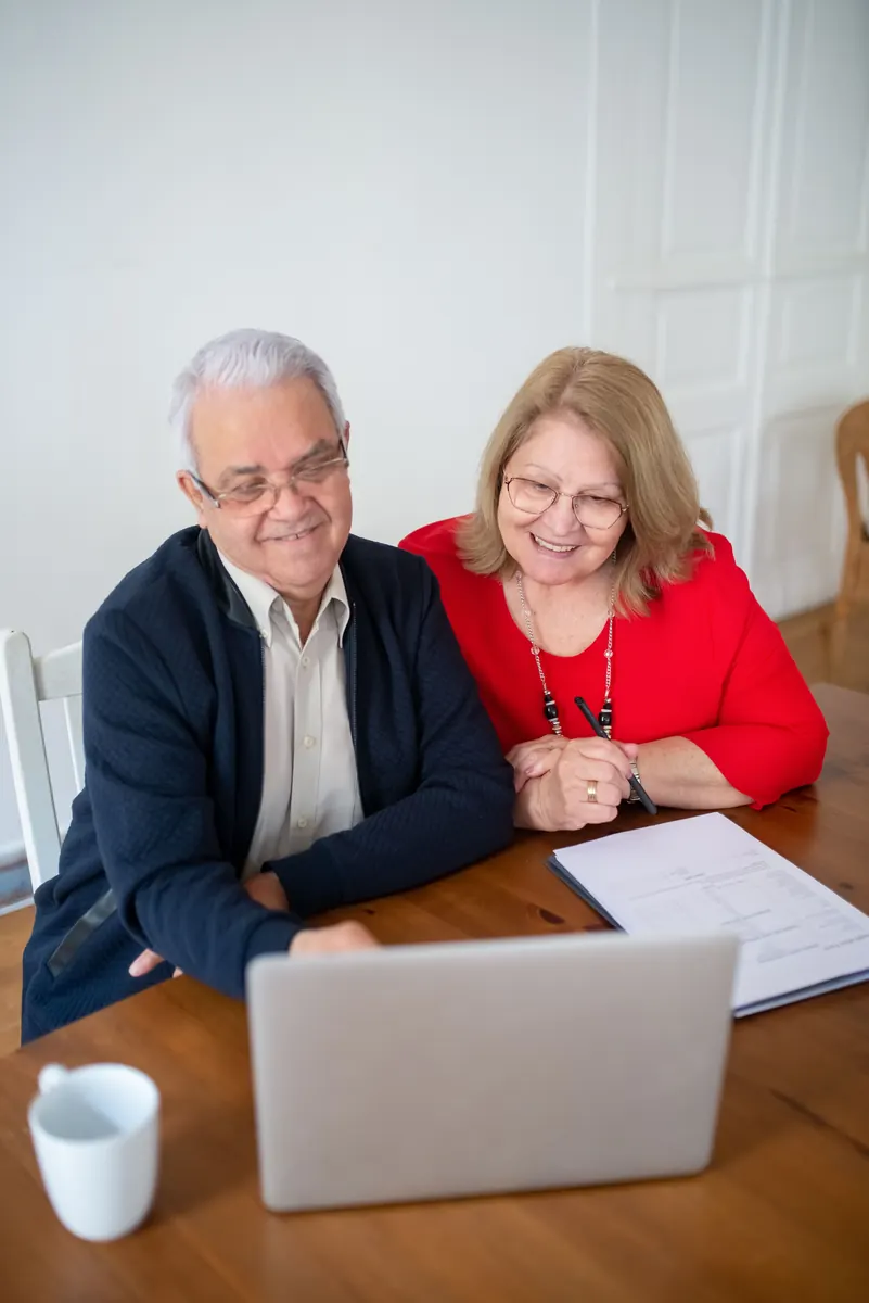A smiling older couple at a wooden table looking at a laptop, with papers and a mug nearby