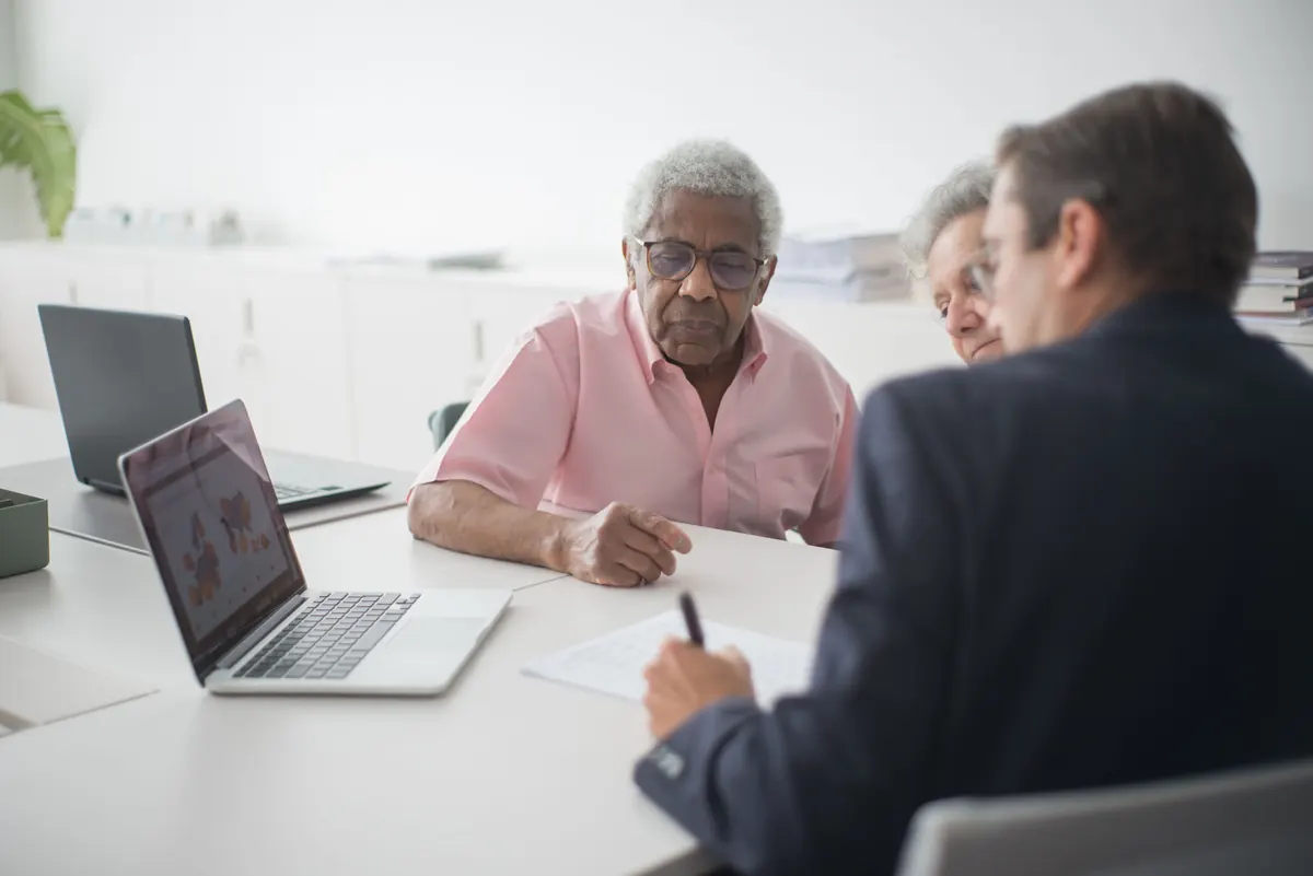 An older couple at an office table with a man in a suit who is taking notes, laptops nearby