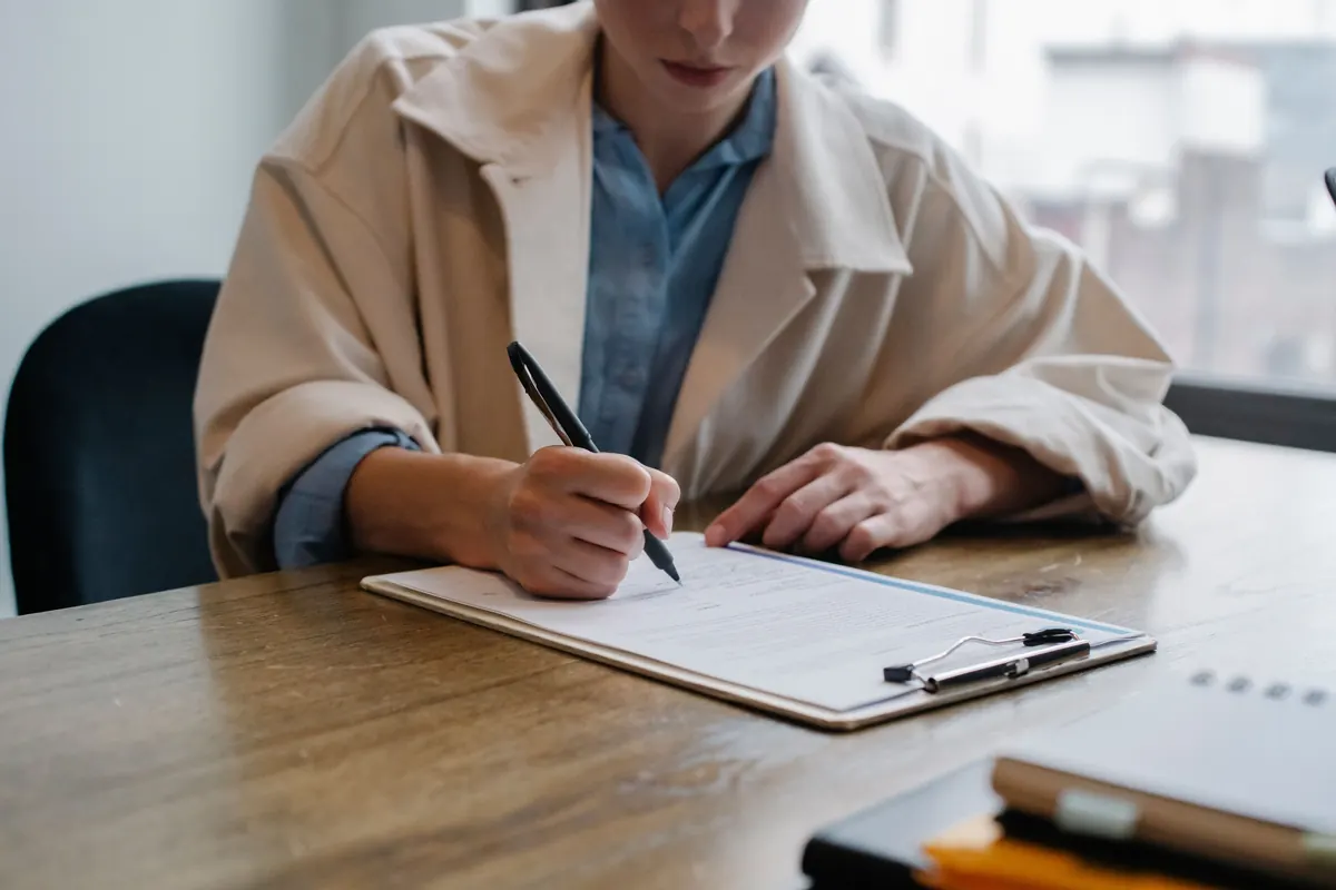 A woman in a beige coat writing on a form on a clipboard at a wooden table by a window