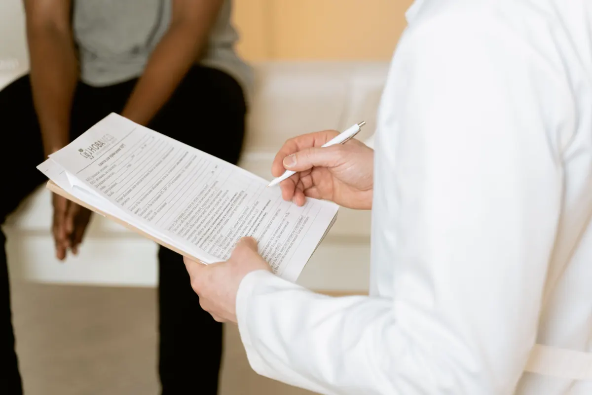A person in a white coat holding a pen and a form on a clipboard while a seated person waits nearby