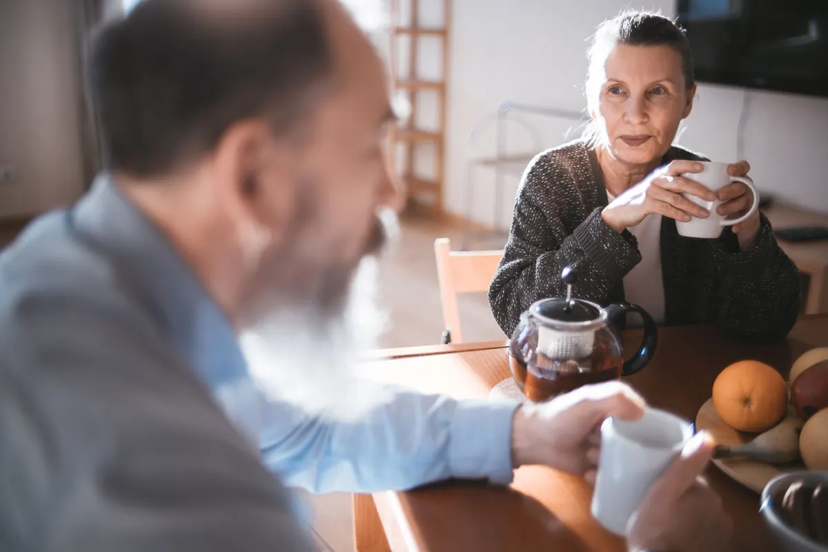 An older woman holding a mug at a kitchen table with a teapot and fruit, a bearded man in the foreground