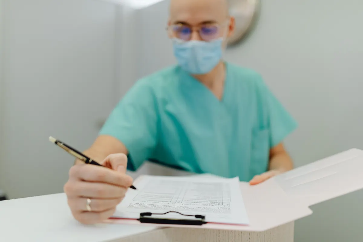 A health worker in green scrubs, a face mask and glasses writing on a clipboard at a front desk