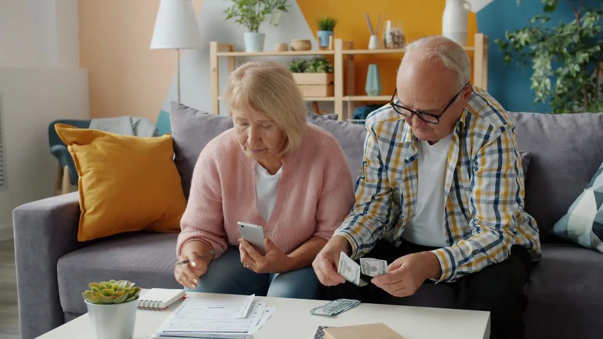 An older couple on a sofa going over paperwork, the woman holding a phone and the man counting cash