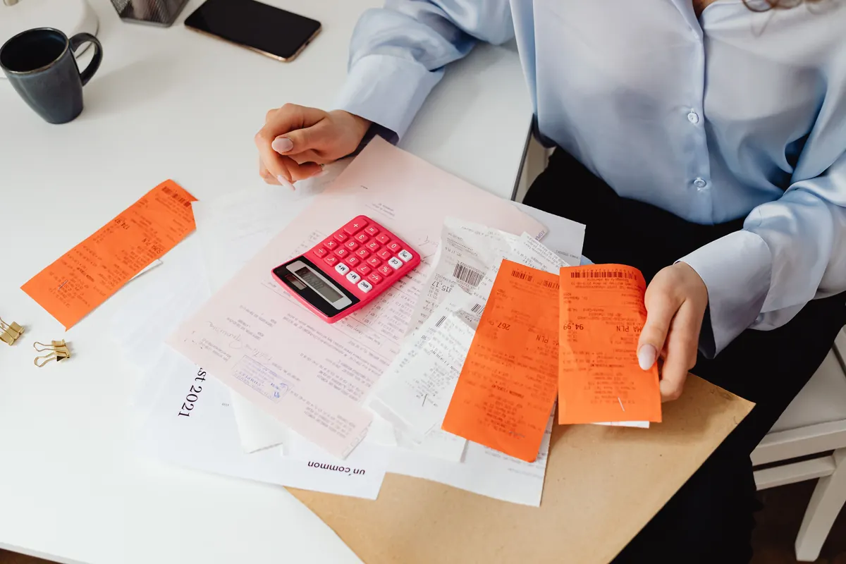 A woman sorting orange receipts and papers with a pink calculator on a white desk
