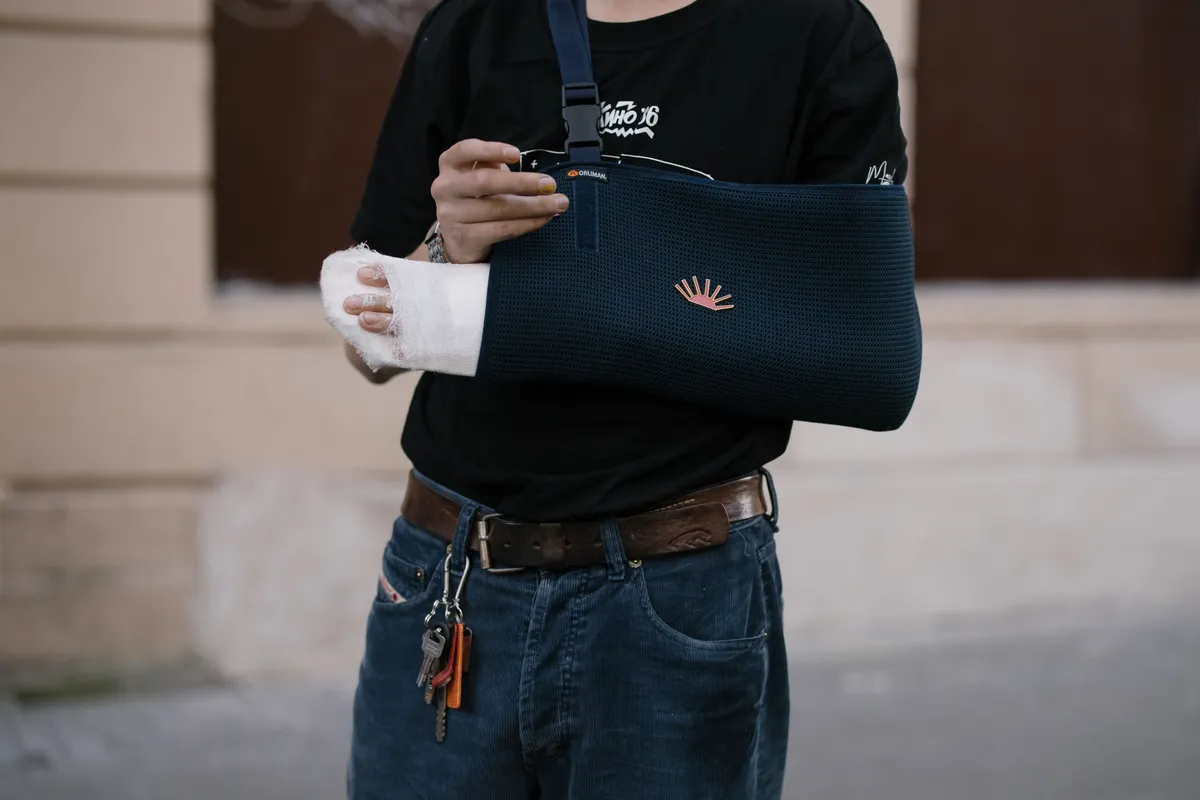 A person in a black T-shirt and jeans with a bandaged hand and forearm resting in a navy arm sling