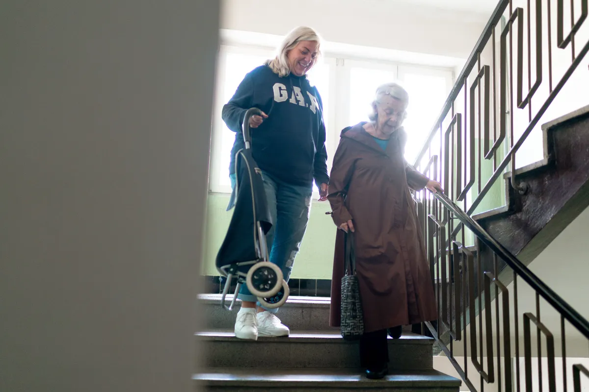A smiling woman with a rolling shopping cart walking down the stairs beside an older woman holding the handrail