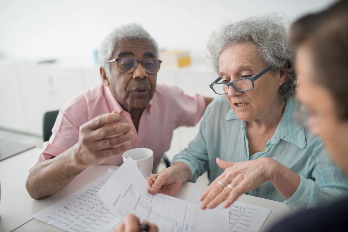 An older man and an older woman with glasses talking over printed forms at a table with a third person